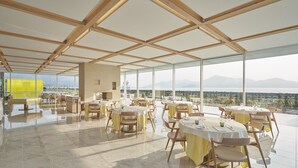 A spacious dining area with high ceilings and large windows offering a mountain view. Tables are set with yellow tablecloths and chairs. - Simose Art Garden Villa (Otake)
