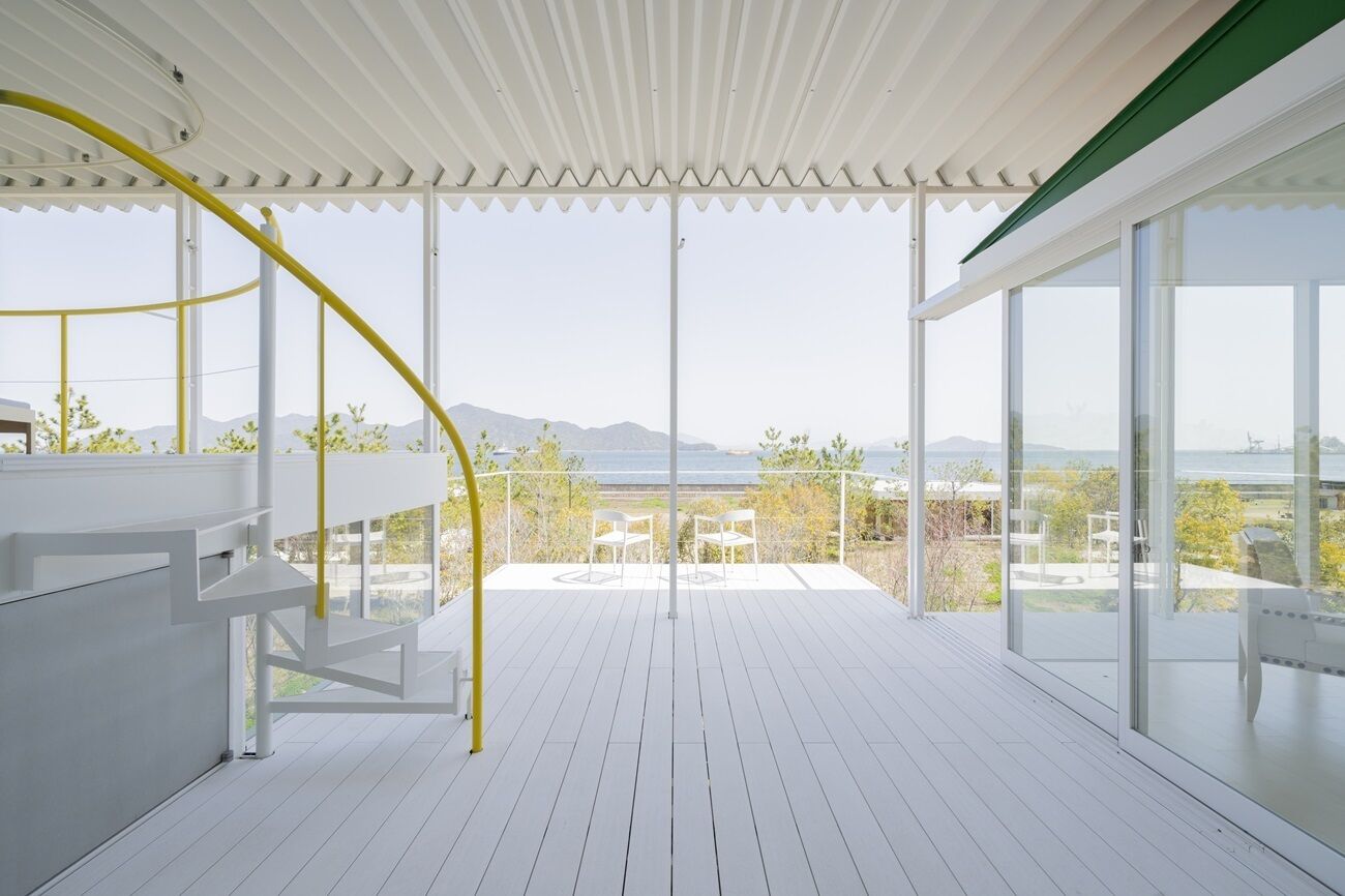 Un balcon spacieux offrant une vue sur la mer et les montagnes, avec des rampes blanches et un escalier en colimaçon jaune.