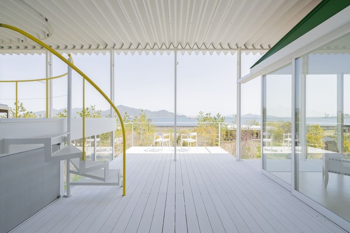 A spacious balcony with a view of the sea and mountains, white railings, and a yellow spiral staircase.