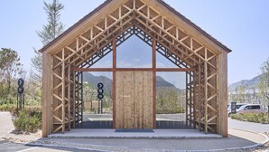A wooden structure with a triangular roof and large glass windows, located at a roadside with a clear sky and mountains in the background. - Simose Art Garden Villa (Otake)