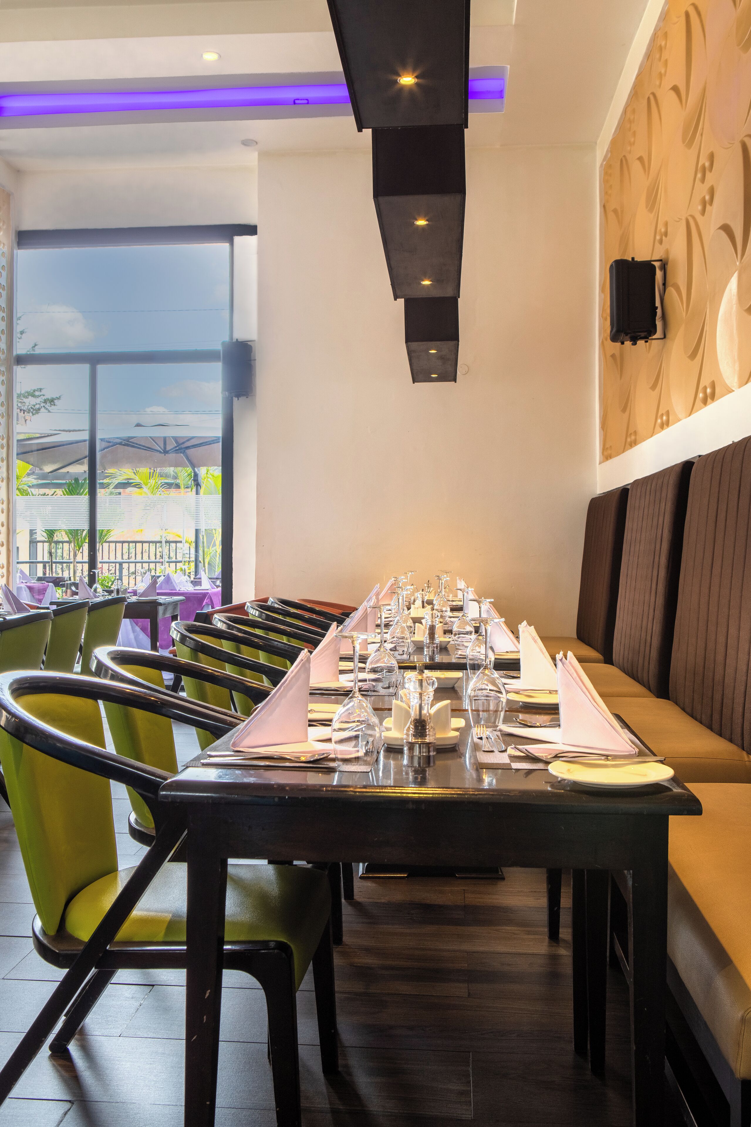 Un ristorante con un lungo tavolo apparecchiato per la cena, sedie verdi e vista attraverso ampie finestre.