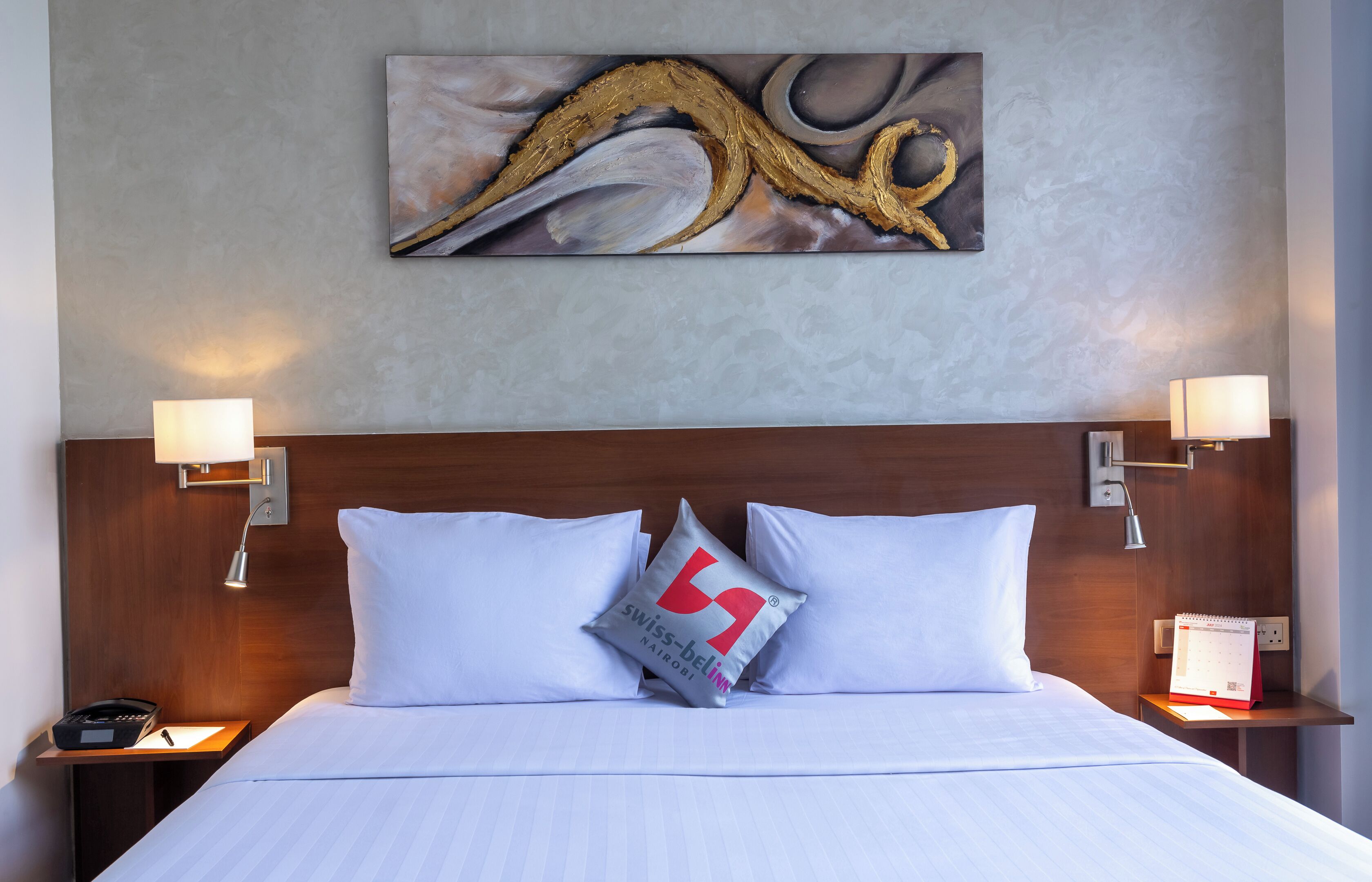 Une chambre d’hôtel avec un grand lit, deux tables de chevet, une petite table d’appoint, une œuvre d’art accrochée au mur et une lampe murale.