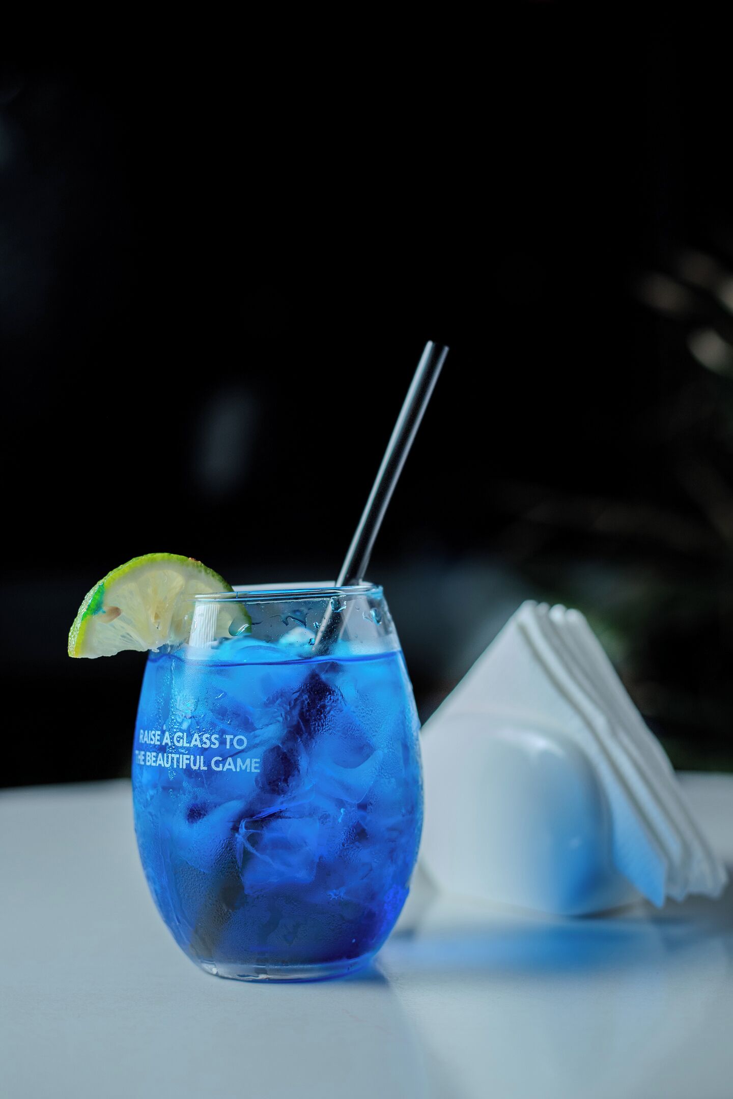 Un vaso de bebida azul con un gajo de lima y una pajita, acompañado de una servilleta y una taza blanca.