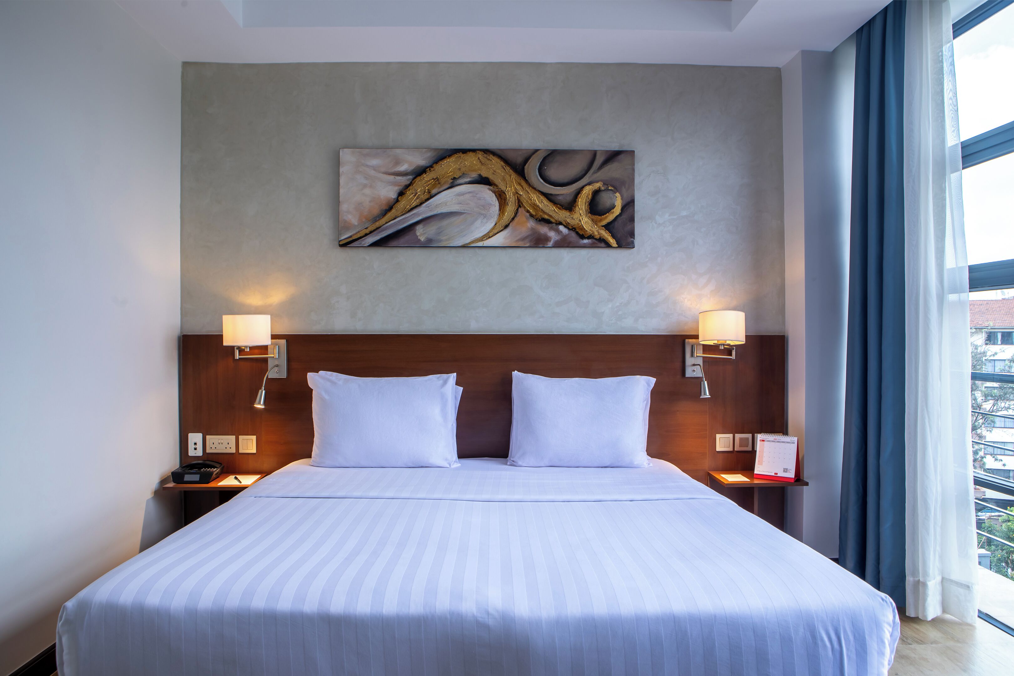 A hotel room with a large bed, two bedside lamps, a wall art piece, and a window with curtains.