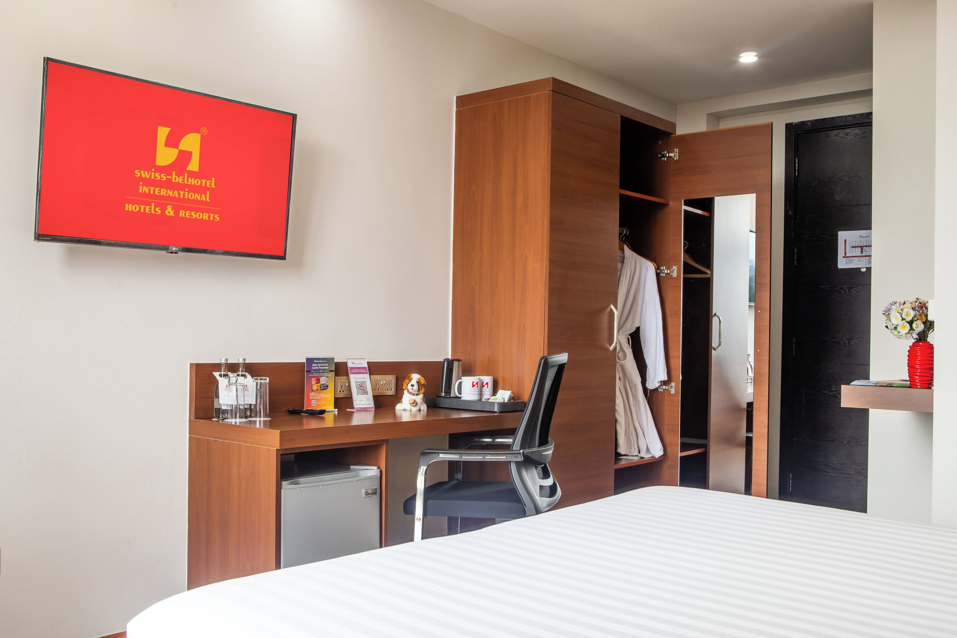 A hotel room with a bed, a desk with a chair, a wardrobe, a small fridge, and a TV displaying the hotel’s name.