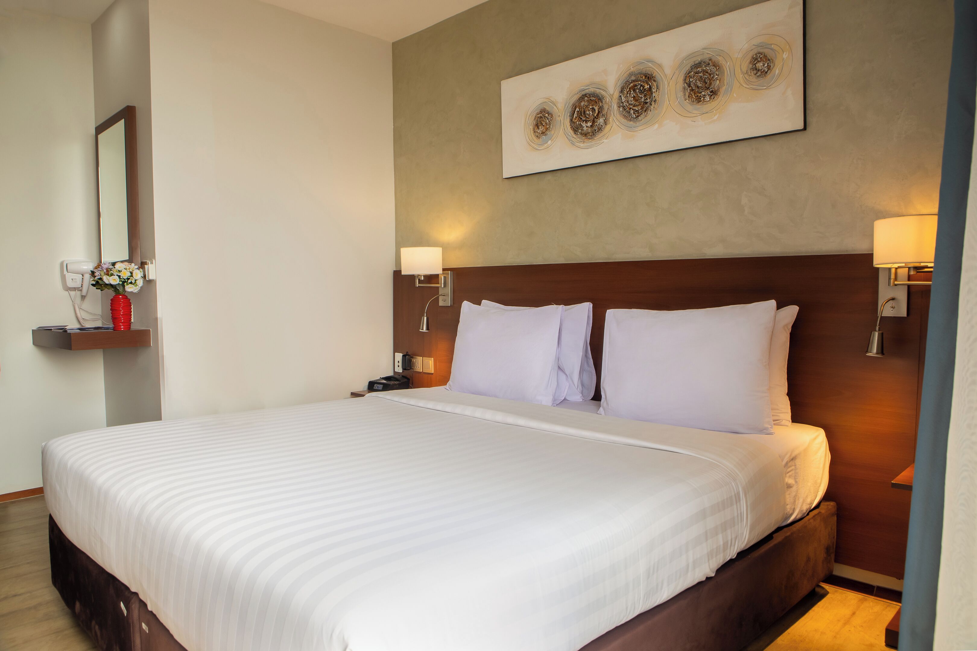 A hotel room with a large bed, bedside lamps, a wall-mounted artwork, and a bedside table with a vase.