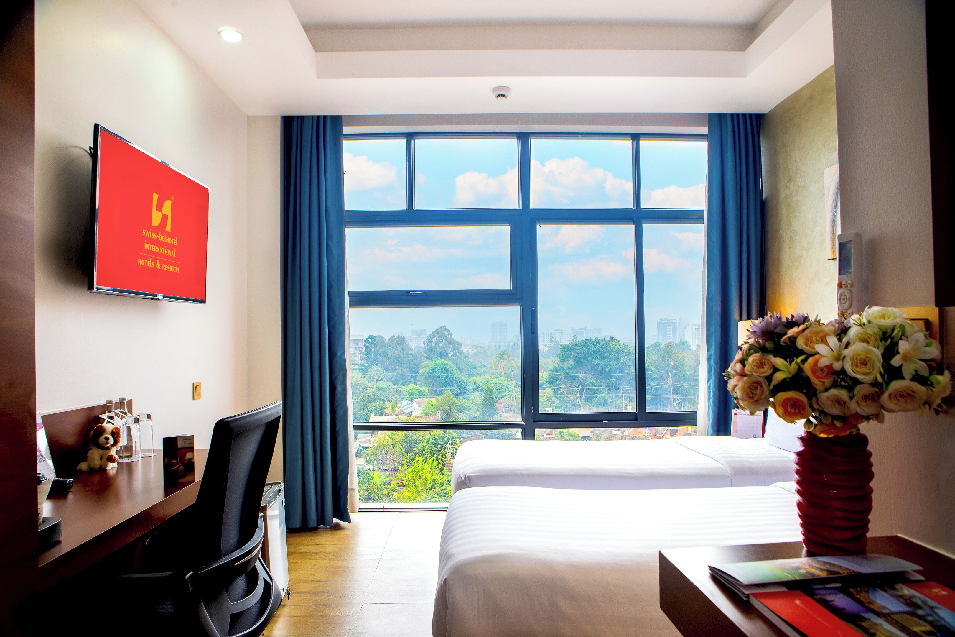 A hotel room with a bed, a desk with a computer, a TV, and a large window with a view of the city.