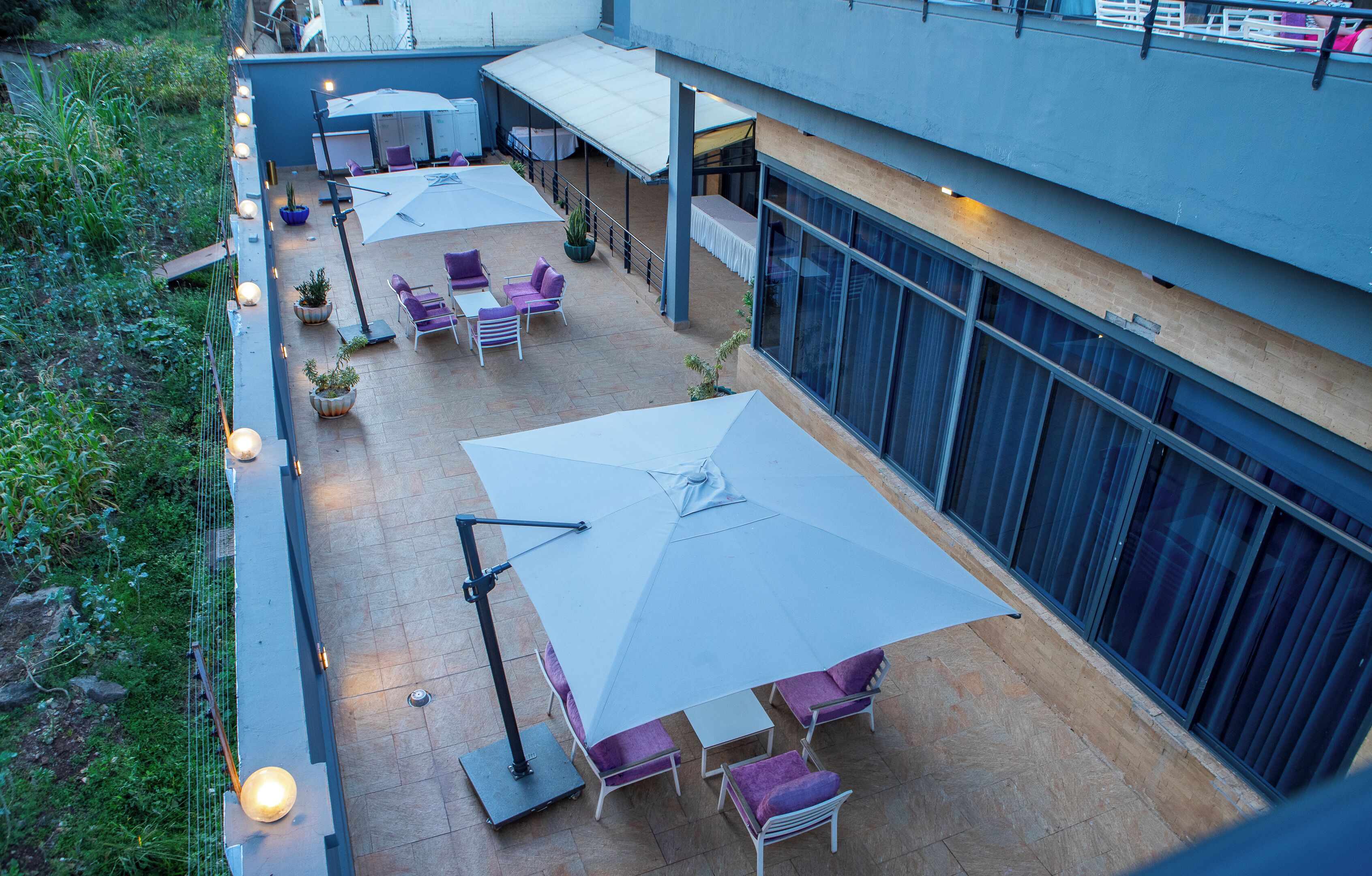 Een terras met meerdere tafels en stoelen onder grote parasols, omgeven door groen en een modern gevelontwerp.