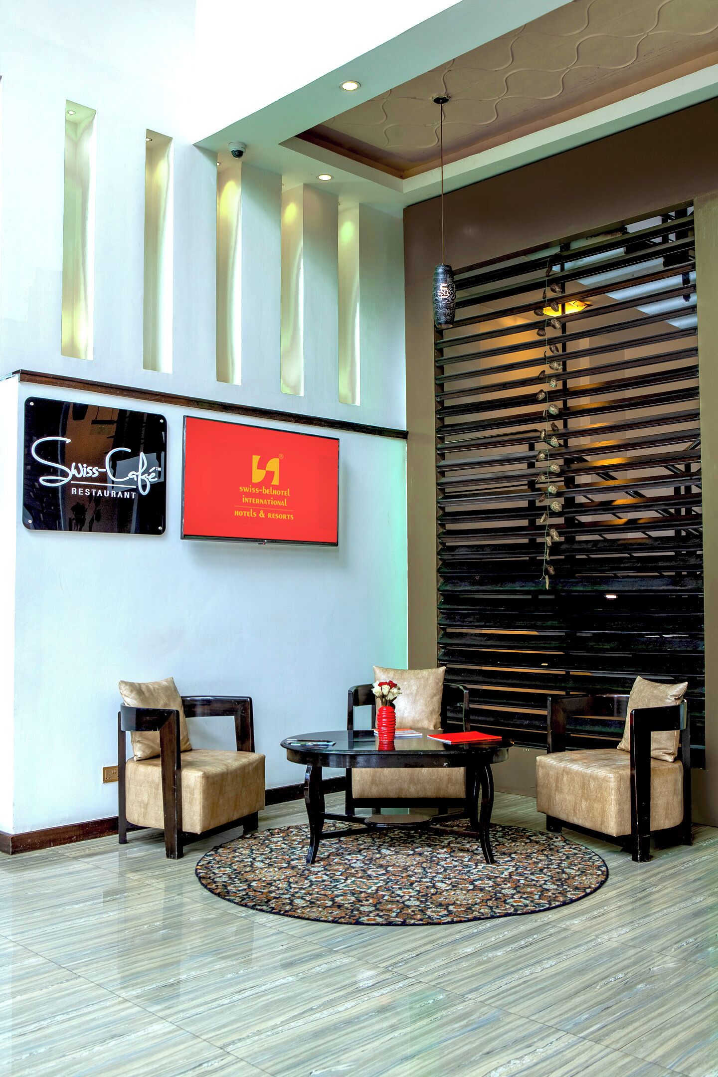 Een lobby met een receptiebalie, twee fauteuils, een bijzettafeltje, een kamerbreed tapijt en een muur met een bord voor Swiss-Café en een groot scherm.