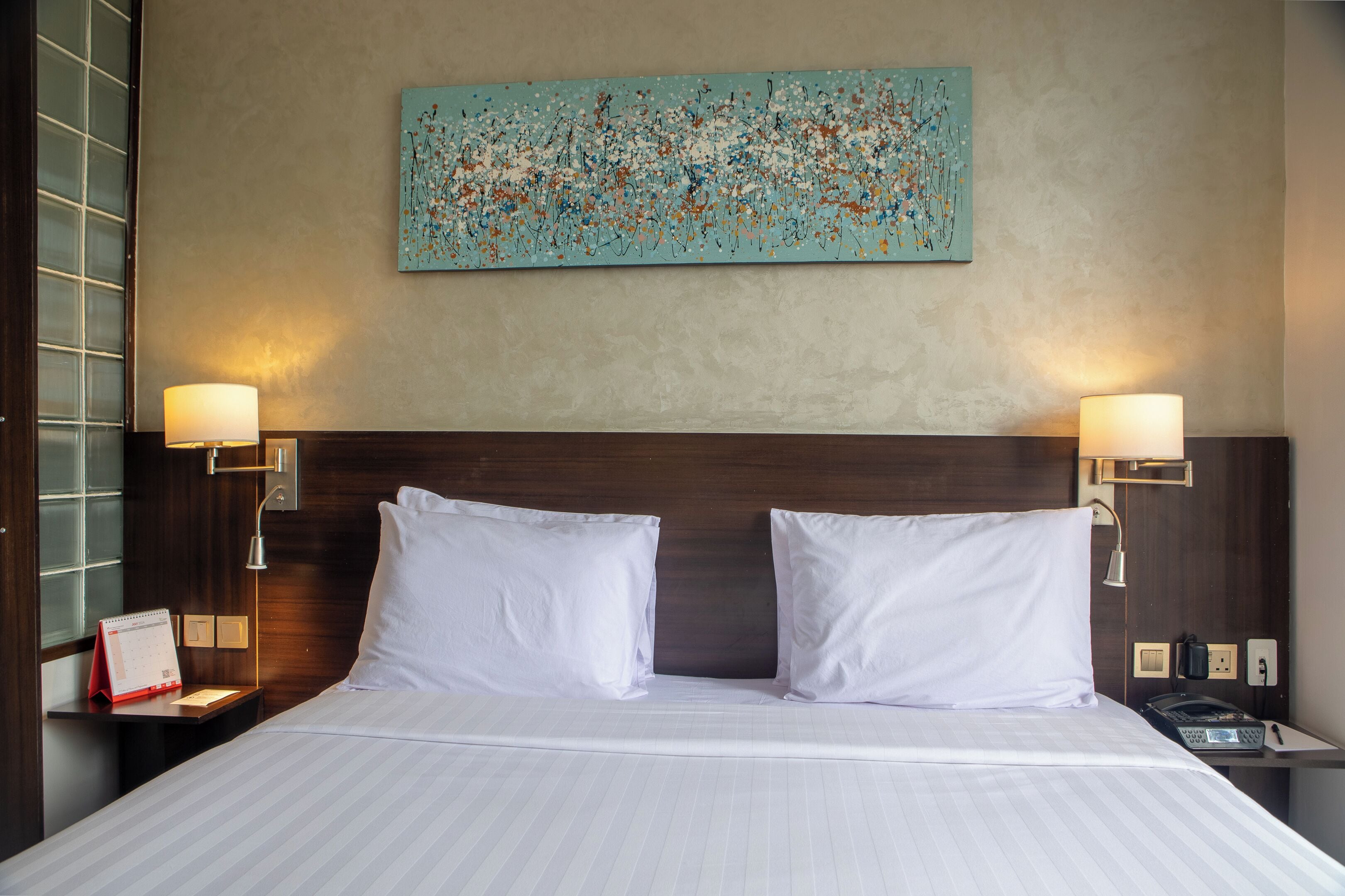 Quarto de hotel com uma cama grande, duas luminárias de cabeceira, um relógio de parede e um calendário na mesinha de apoio.