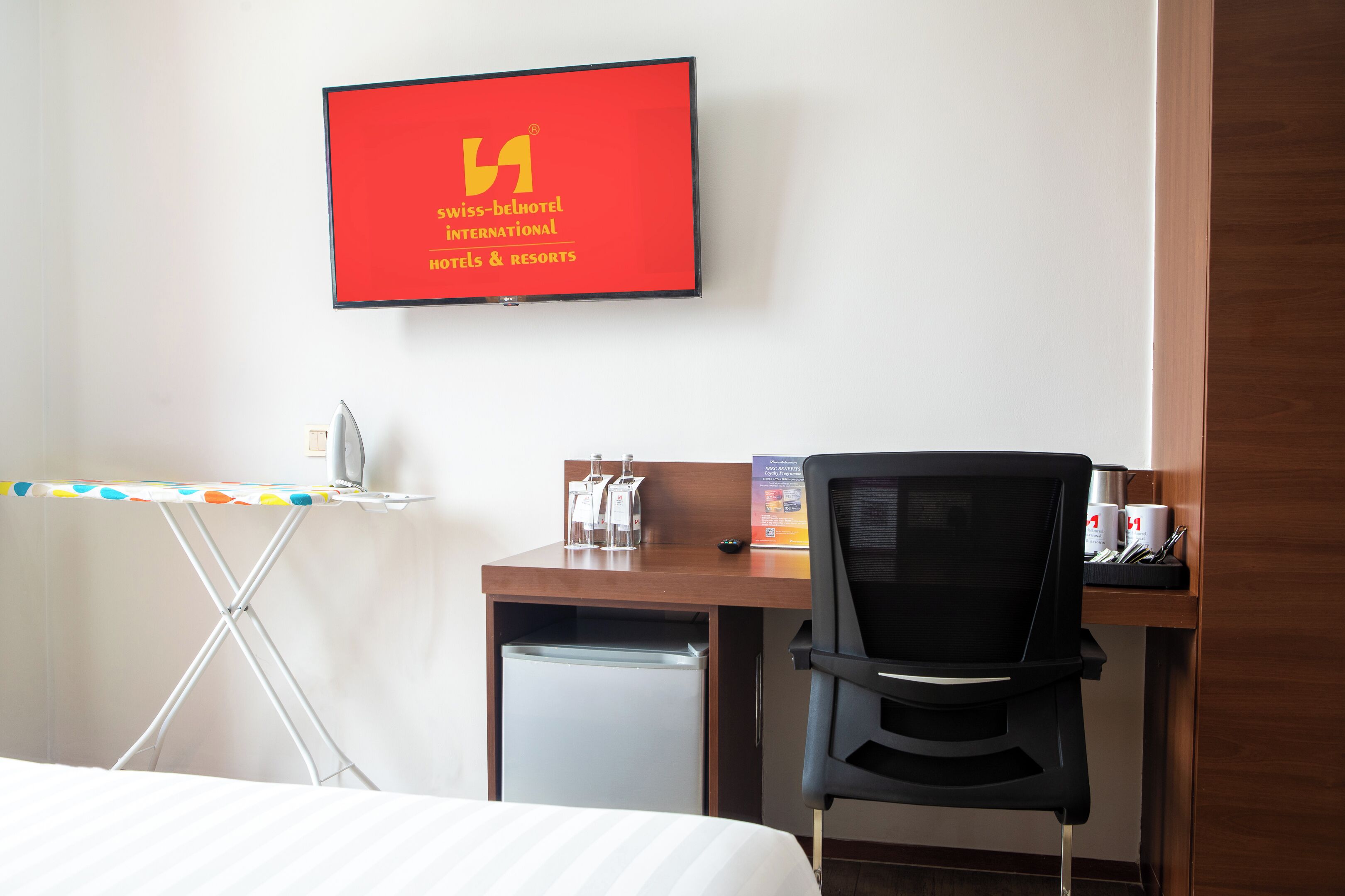 A hotel room with a bed, a desk, a chair, a TV mounted on the wall, and a small refrigerator.