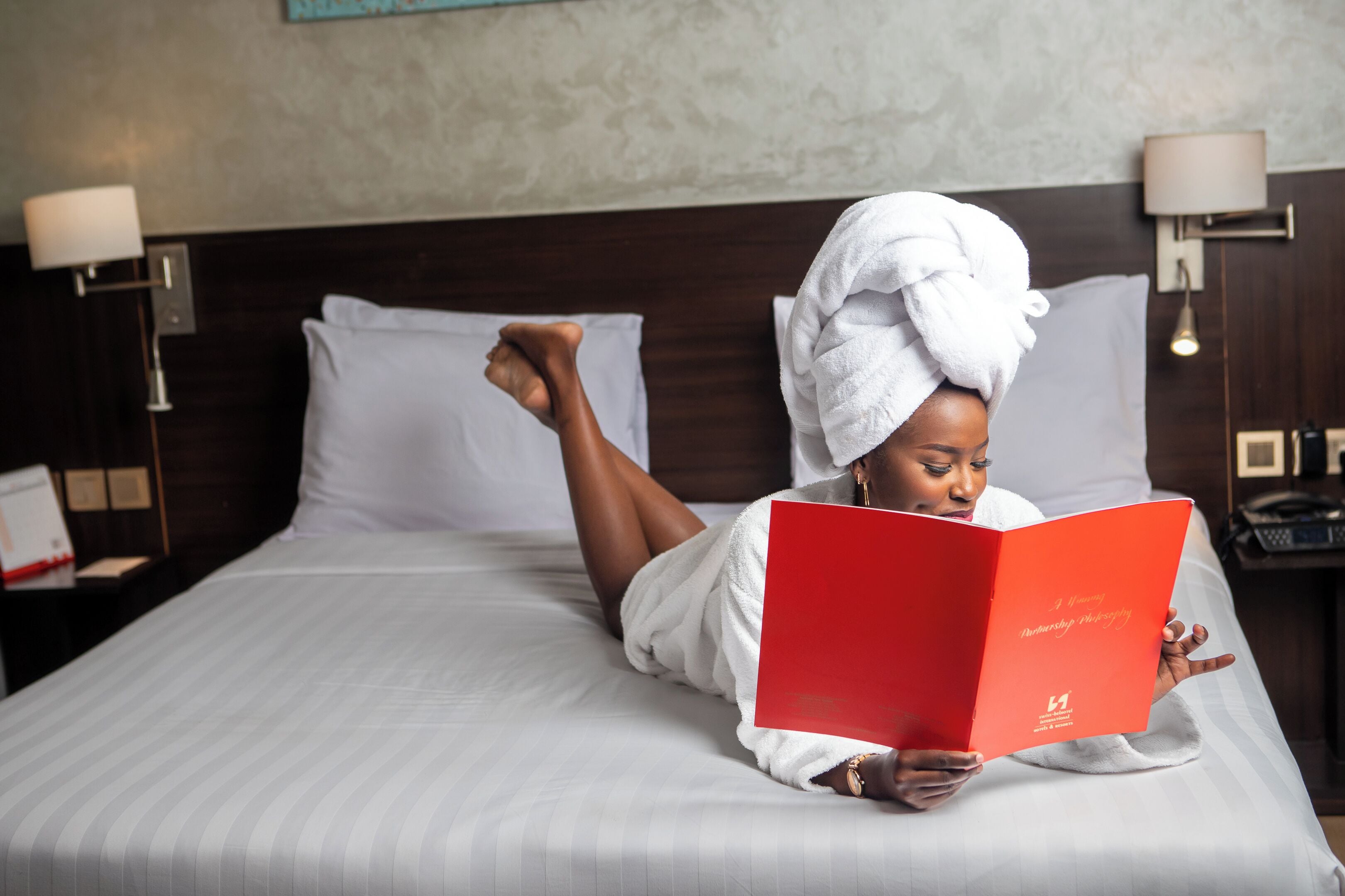 A person lying on a bed with a towel wrapped around their head, reading a red book.