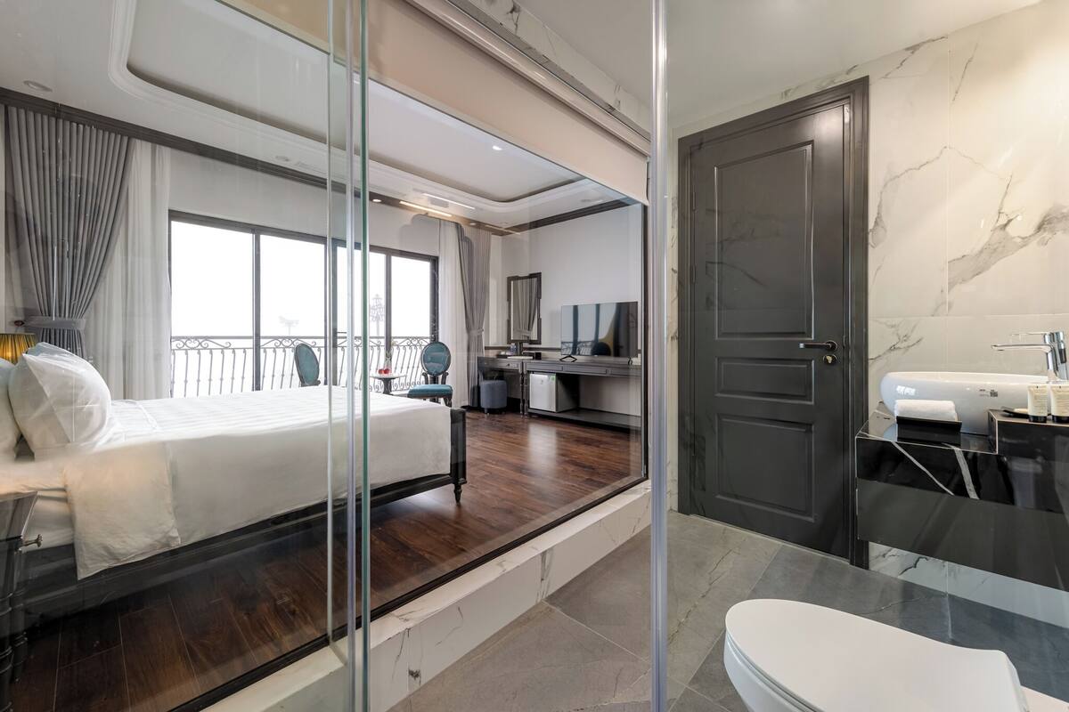 Una habitación de hotel moderna con una cama grande, un balcón, un escritorio, un baño con espejo y lavabo, y un piso de mármol.