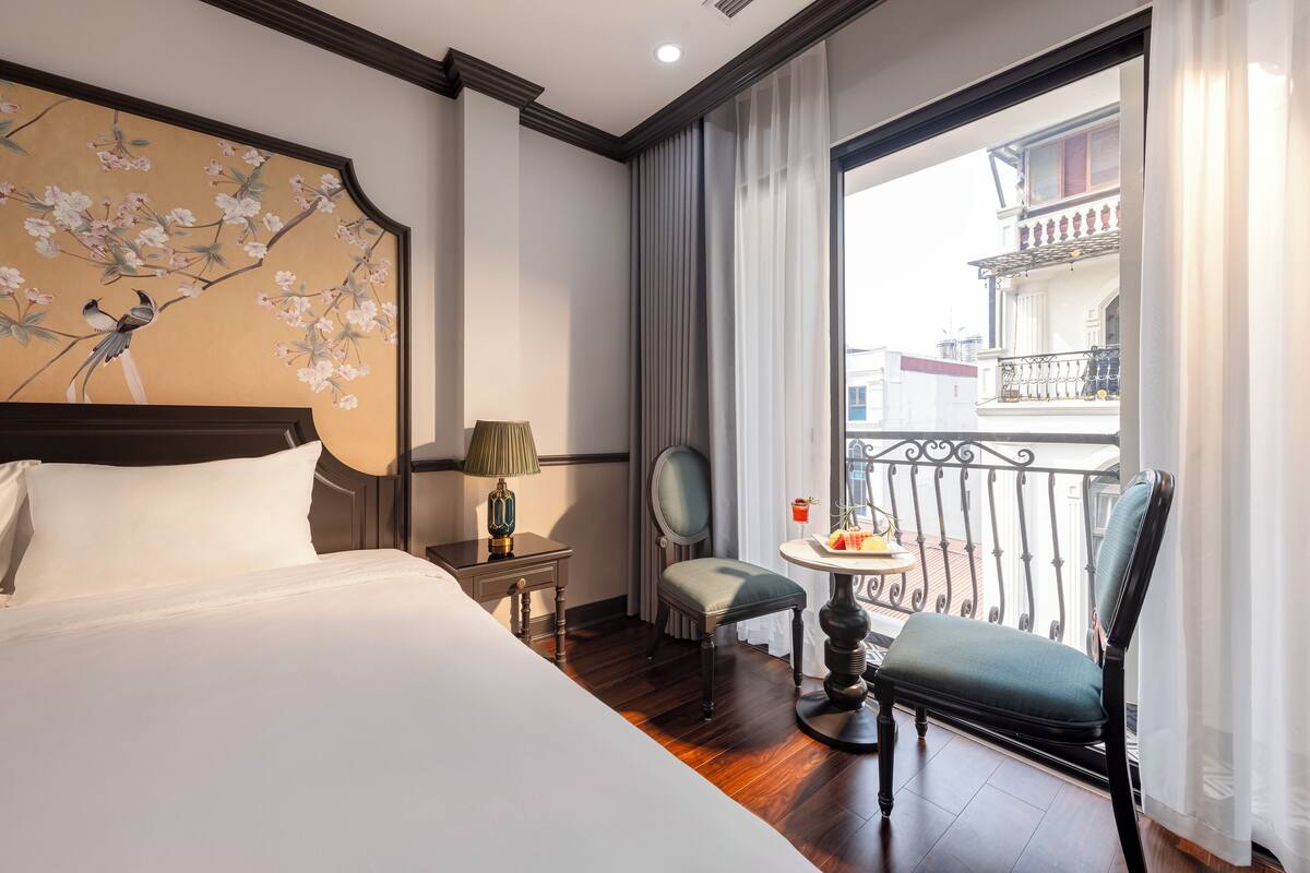 Habitación de hotel con una cama grande, una silla, una mesita con fruta y un balcón con barandilla.