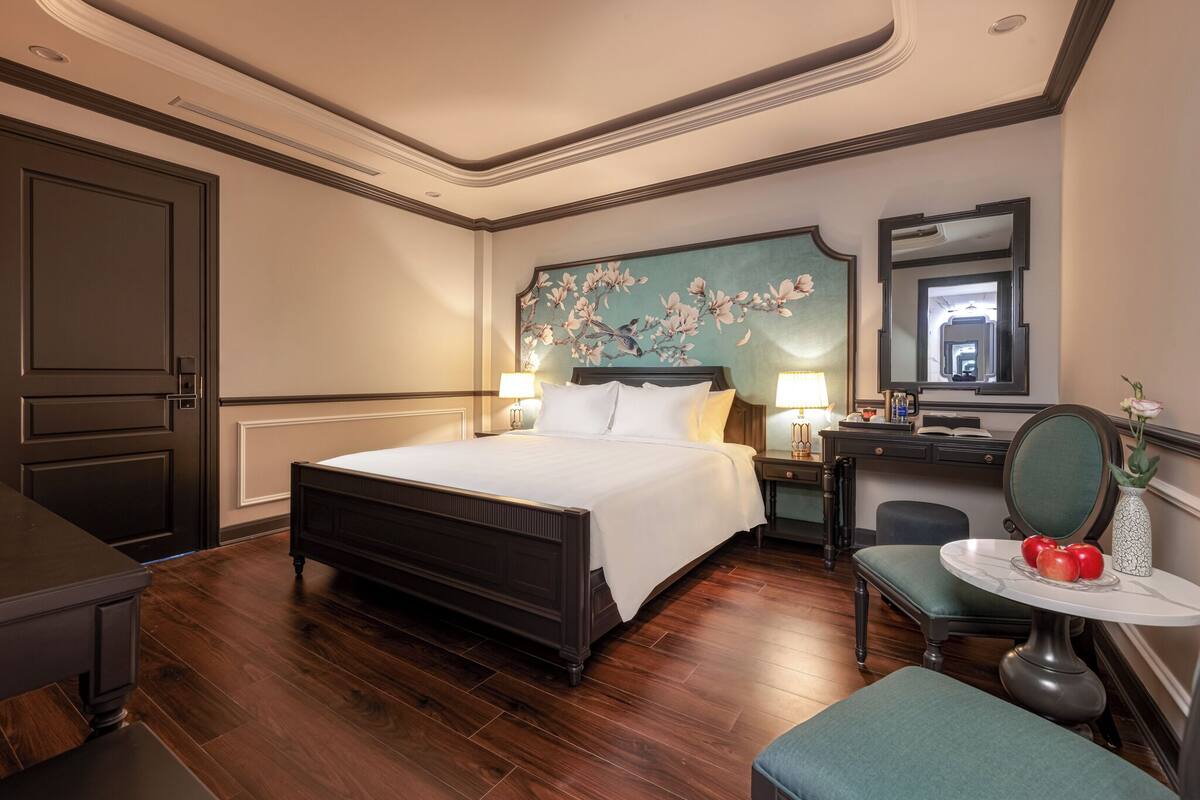 Habitación de hotel con una cama grande, un escritorio con silla, una mesita con manzanas y un espejo.