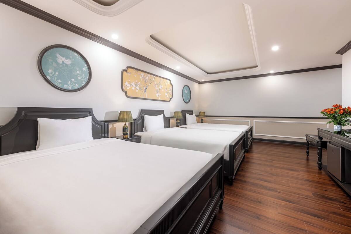 Habitación de hotel con tres camas individuales, suelo de madera, un reloj y una obra de arte floral en la pared.