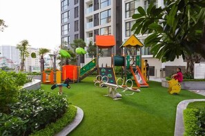 A playground with various play structures, surrounded by greenery and a building in the background.