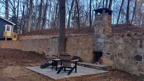 Une cheminée en pierre avec un conduit de cheminée, entourée d’un mur de pierre et d’une terrasse en bois agrémentée de fauteuils Adirondack.