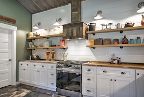 Una cocina con gabinetes blancos, estufa de acero inoxidable y repisas de madera con diversos objetos de cocina.