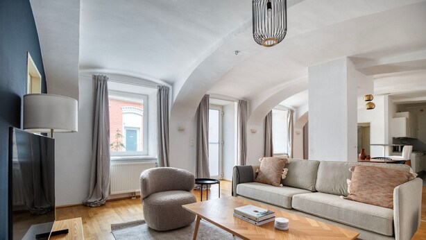 A modern living room with a sofa, armchair, and coffee table, large windows with curtains, and a television.