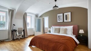 A bedroom with a large bed, a desk, and a chair. There are two windows with curtains, a radiator, and a ceiling fan.
