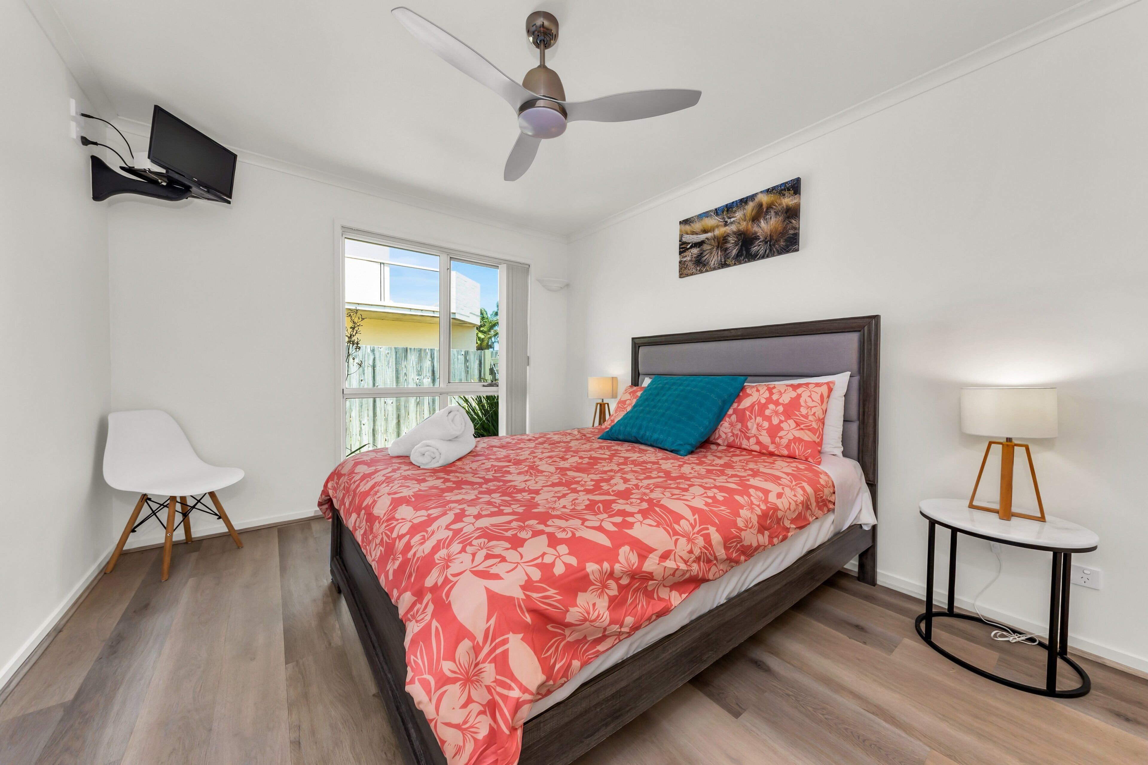 Une chambre à coucher comprenant un lit, un ventilateur de plafond, un téléviseur fixé au mur, une fenêtre et une petite table avec une lampe.