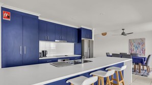 Una cocina moderna con armarios azules, encimera blanca y refrigerador de acero inoxidable. Se observan tres taburetes blancos tipo barra y una mesa de comedor con sillas al fondo.