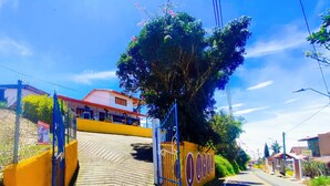 A paved pathway leading to a gated entrance with a blue gate, a yellow wall, and a building with a red roof in the background. - JUHALU (Jericó)