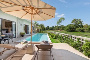 A poolside area with a lounge chair, umbrella, and a dining table with chairs. - Sereniwood 2 - 3 Bedroom Pool Villa (Koh Samui)