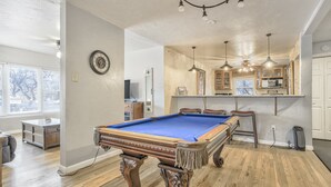 A pool table in a room with wooden flooring, a kitchen area with cabinets and a bar, and a clock on the wall. - The Keys (Colorado Springs)