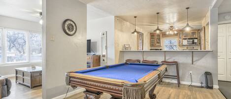 A pool table in a room with wooden flooring, a kitchen area with cabinets and a bar, and a clock on the wall.