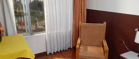 Una habitación con una silla de madera, una mesa amarilla, una ventana con cortinas y vistas al exterior.