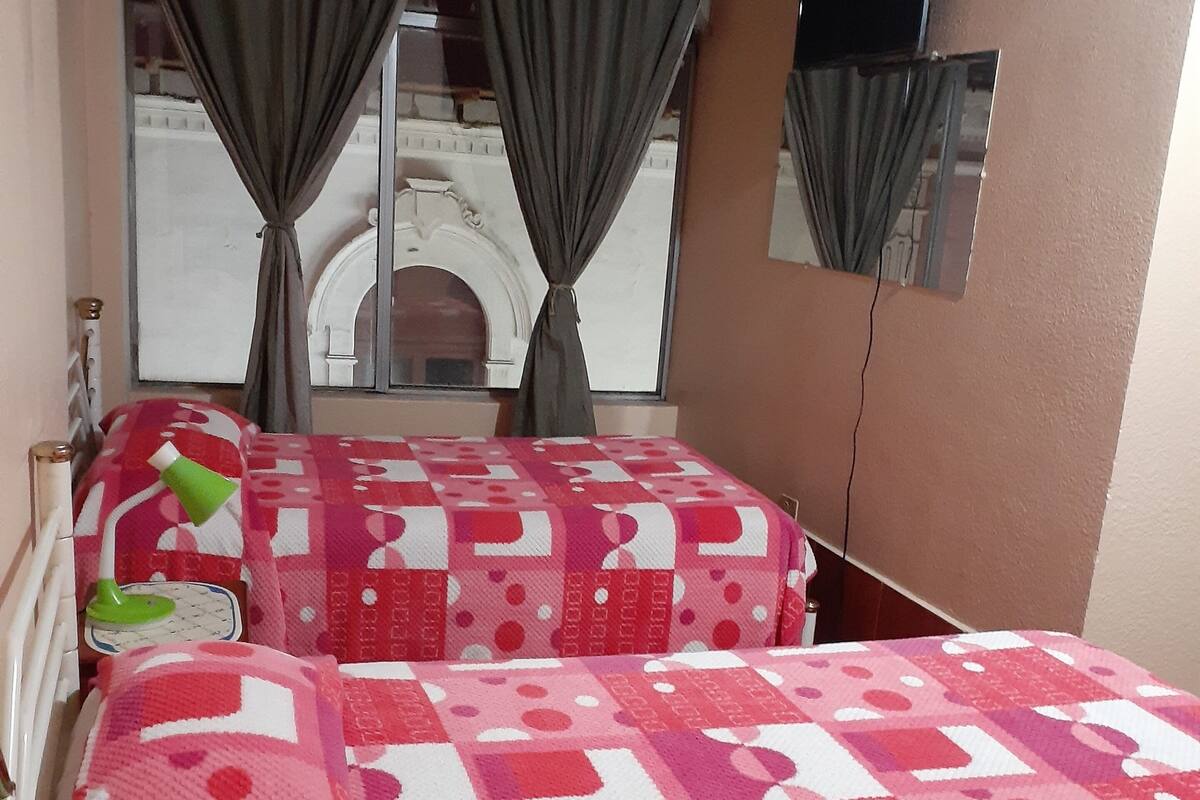 A hotel room with two beds, a window with curtains, a small table with a lamp, and a mirror on the wall.