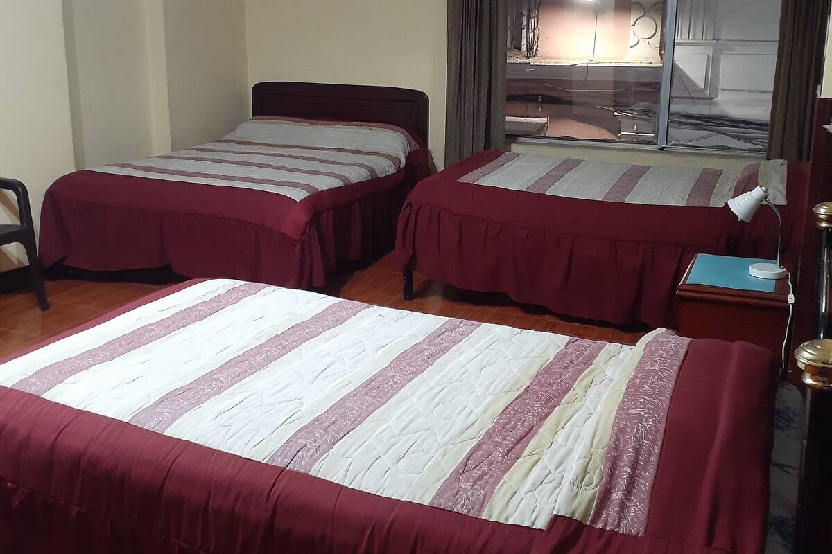A hotel room with two beds, a desk with a lamp, a window with curtains, and a small table with a book.