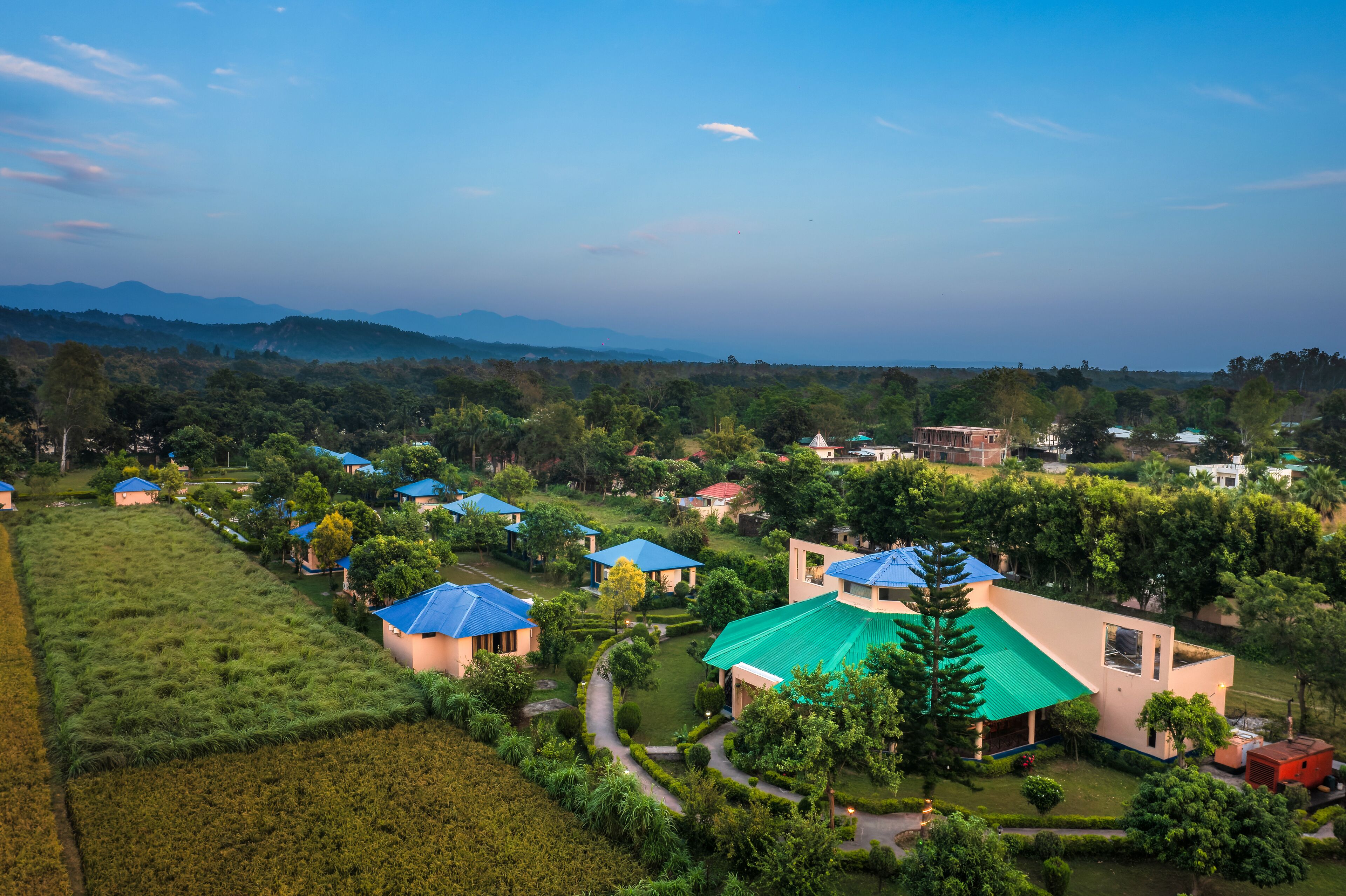 A residential area with multiple houses, some with blue roofs, surrounded by lush greenery and a distant mountain range.