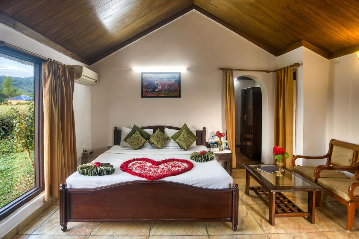 A bedroom with a wooden ceiling, a bed with a heart-shaped decoration, a bedside table with a vase of flowers, and a view of greenery outside.