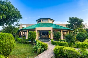 A building with a green roof, surrounded by well-maintained hedges and a stone pathway leading to the entrance. - Tusk And Roar Resort Petfrindly Jungle Cottages (Ramnagar)