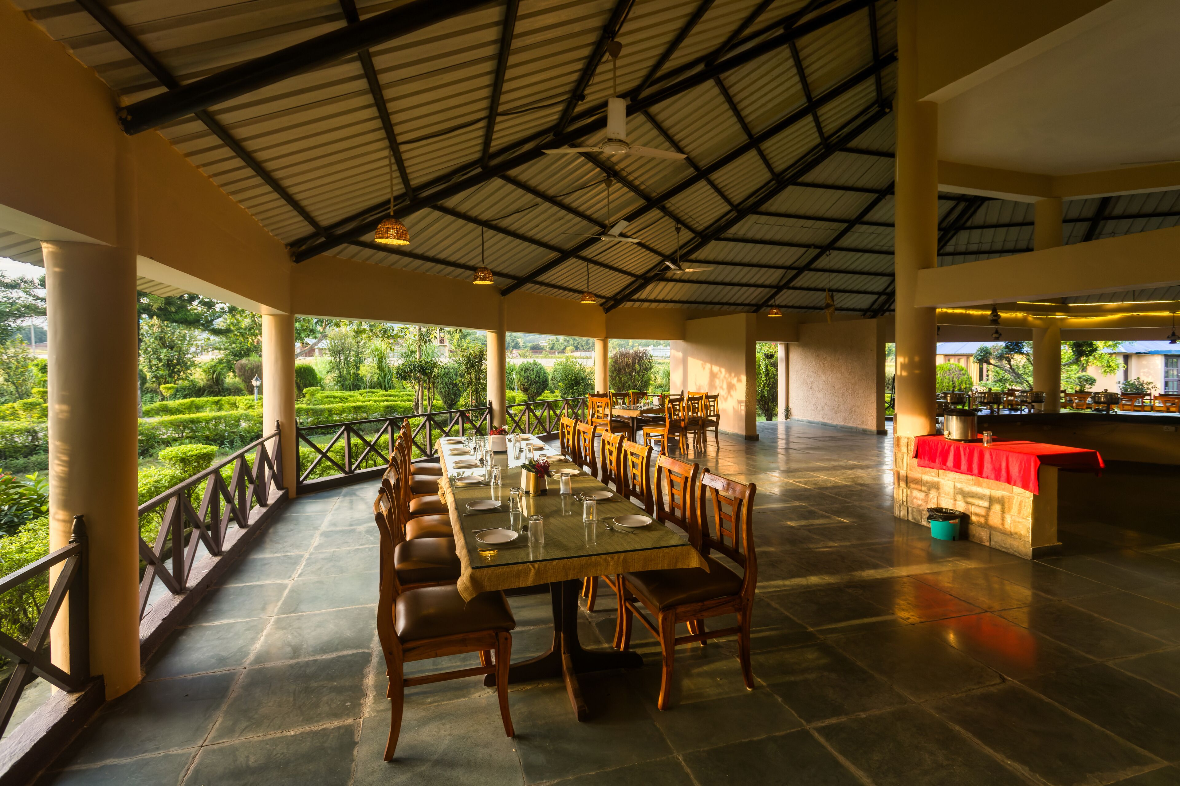 A spacious outdoor dining area with a long table and chairs, surrounded by a railing with a view of a lush garden.