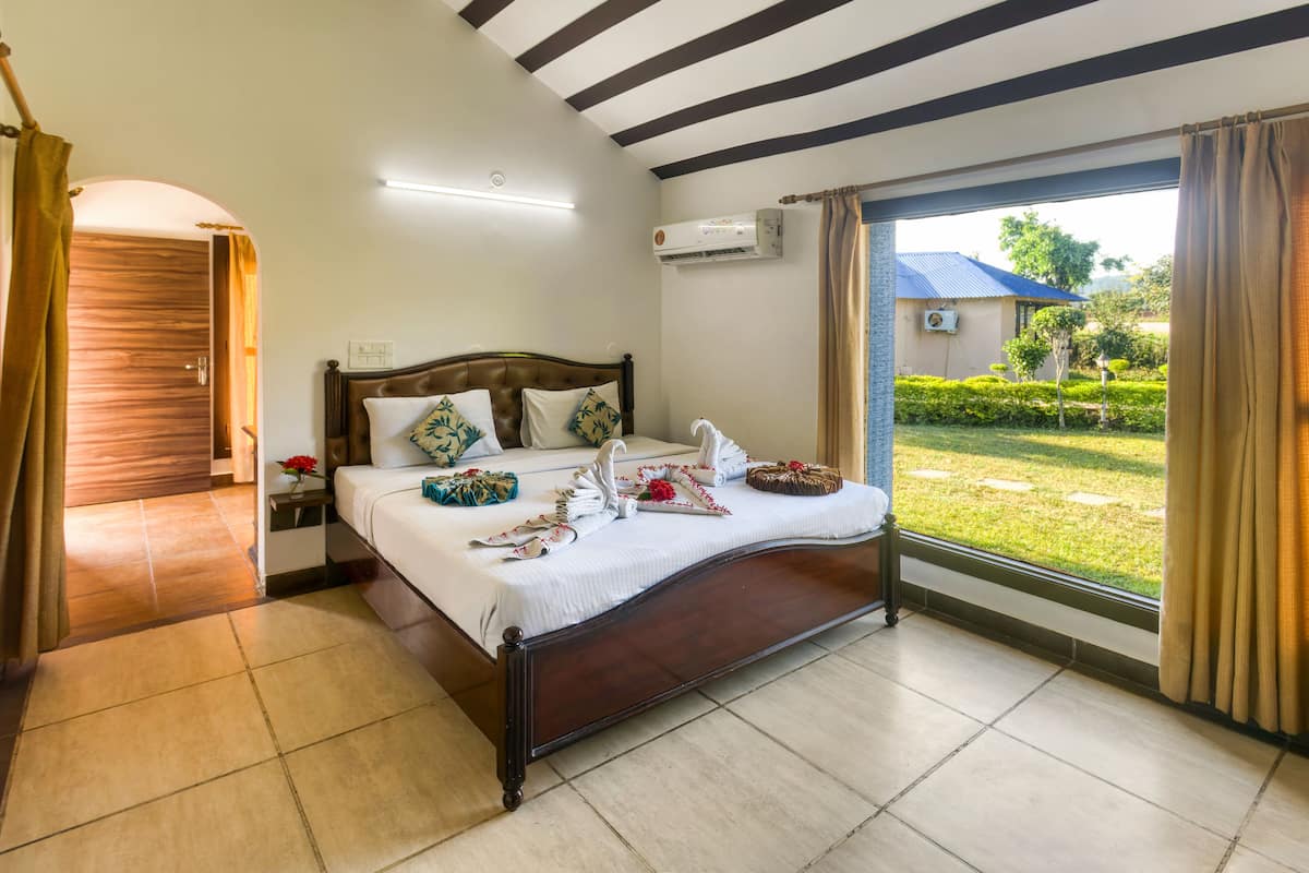 A hotel room with a large bed, a wooden door, a window with curtains, and a view of a garden.