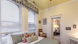 Une chambre à coucher comprenant un lit, deux fenêtres équipées de stores, une chaise, un bureau et une porte donnant sur une autre pièce.