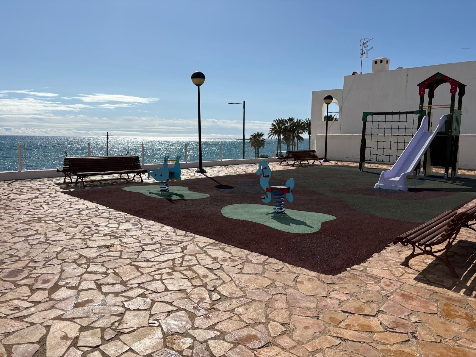 Un parque infantil con tobogán, columpios y un banco junto al mar.