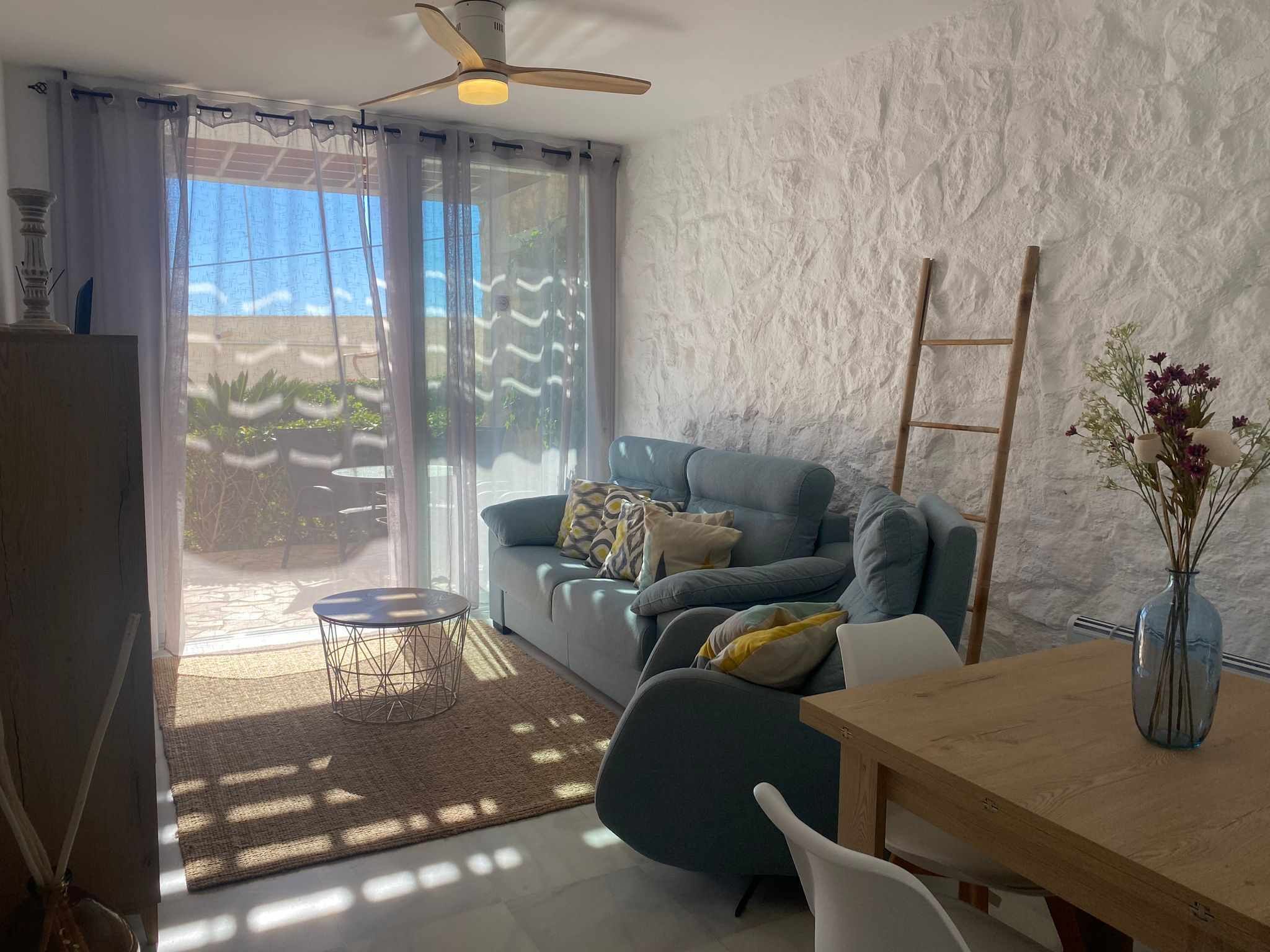 Una sala moderna con un sofá gris, una mesa de comedor de madera y una silla blanca. Hay un ventanal con cortinas translúcidas, un ventilador de techo y una escalera de madera apoyada en la pared.