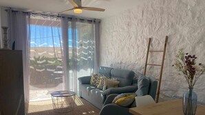 A modern living room with a grey sofa, a wooden dining table, and a white chair. There is a large window with sheer curtains, a ceiling fan, and a wooden ladder leaning against the wall. - Beautiful apartment ideal for families (Mojacar)