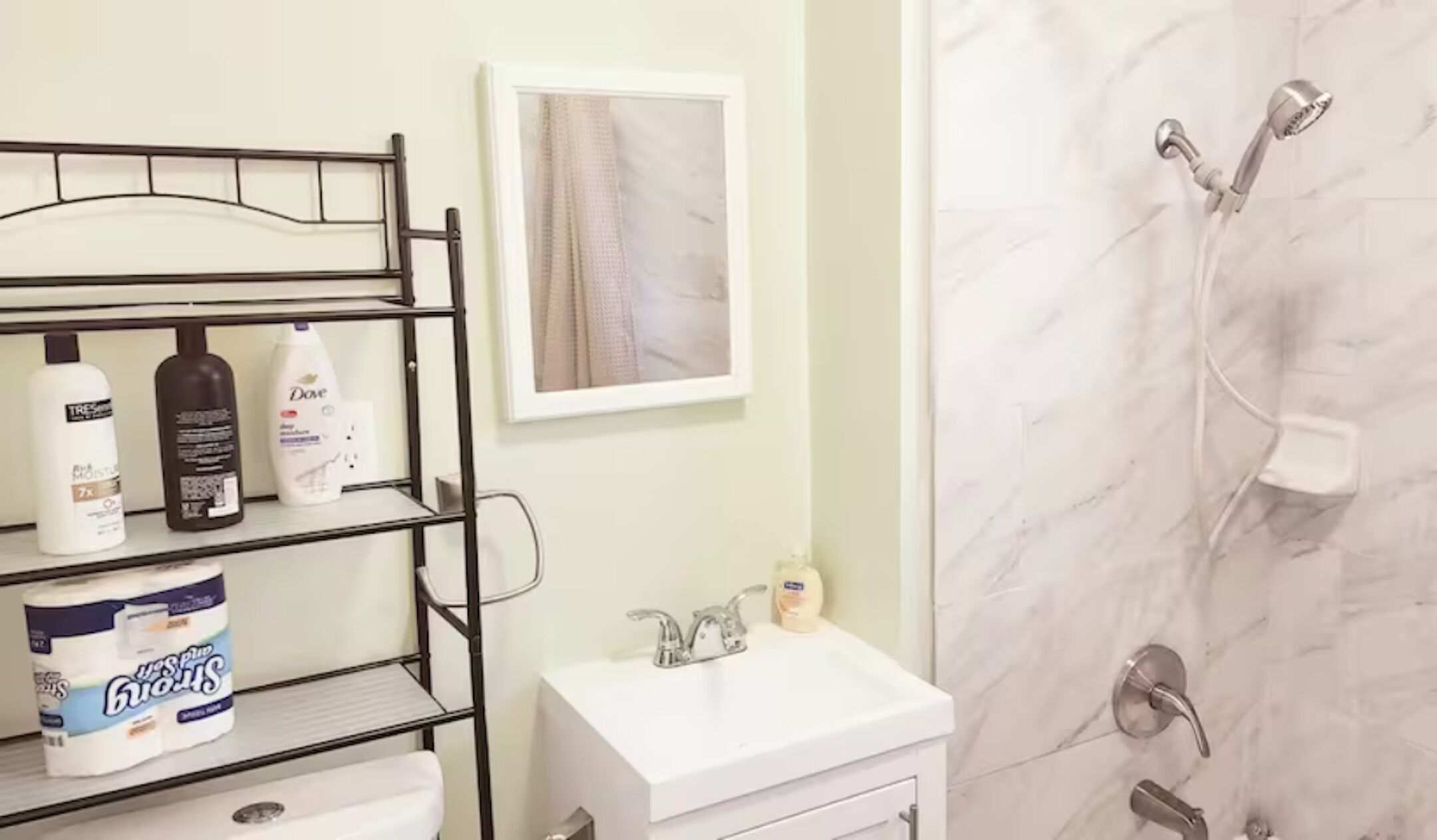 Casa de banho com lavatório branco, chuveiro com chuveiro manual e prateleira com artigos de higiene, incluindo uma embalagem de sabonete Dove e uma caixa de papel higiénico.