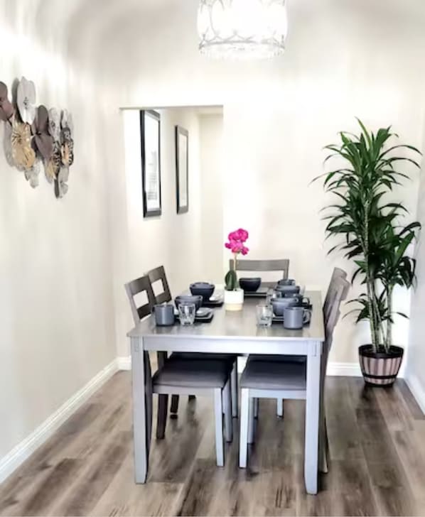 Een eetgedeelte met een tafel voor vier personen, omringd door stoelen. Rechts staat een potplant en boven de tafel hangt een decoratie.