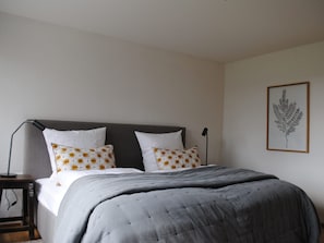 A neatly made bed with a grey duvet, two white pillows with a floral pattern, a framed botanical print on the wall, and a bedside table with a lamp.