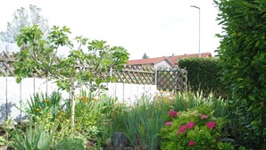 Un jardín con diversas plantas y flores, una cerca de madera y un edificio con tejado rojo al fondo.