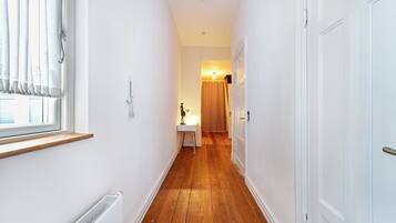 Un pasillo estrecho con suelo de madera, paredes blancas, un radiador, una ventana con cortinas y una puerta al final.