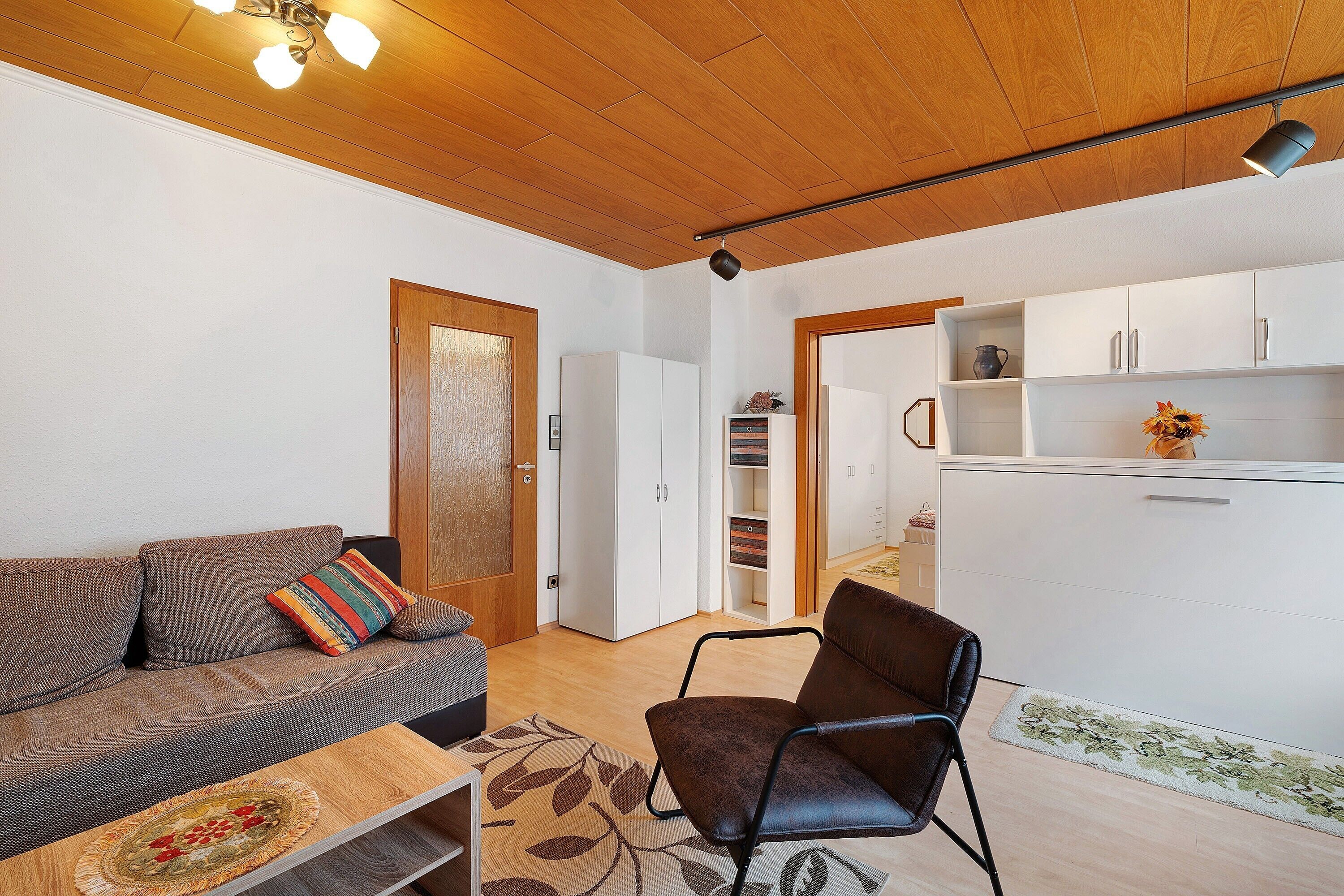 Un soggiorno con un divano, una poltrona, soffitto in legno e un angolo cottura con pensili.