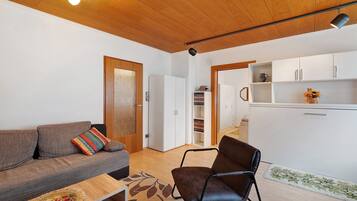 Un soggiorno con un divano, una poltrona, soffitto in legno e un angolo cottura con pensili.